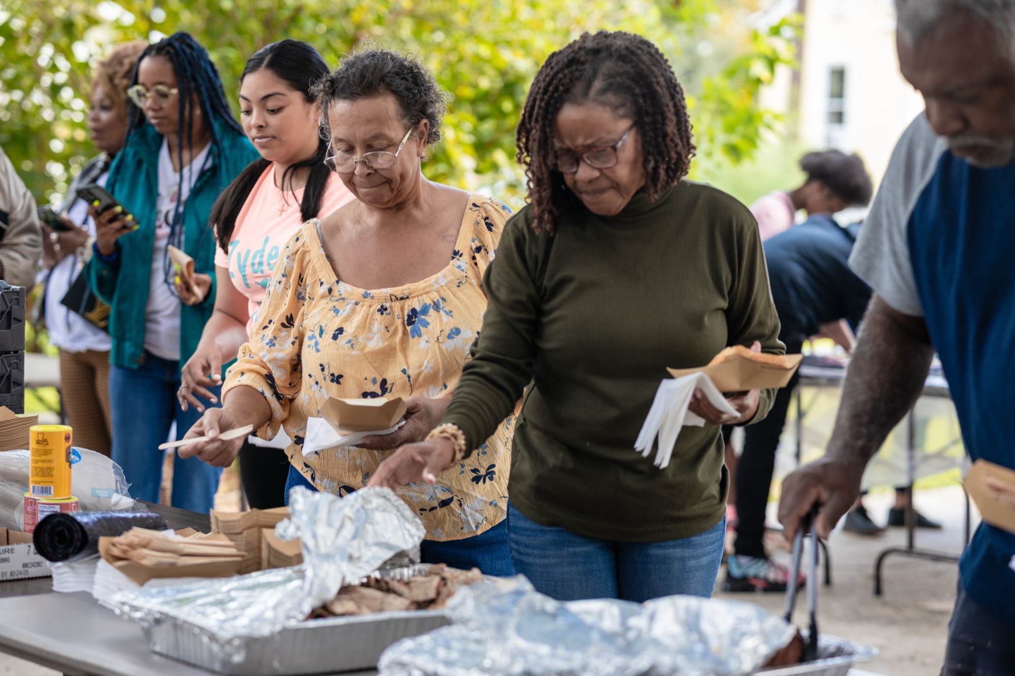 People serving black-pot Creole food out of foil trays at a table